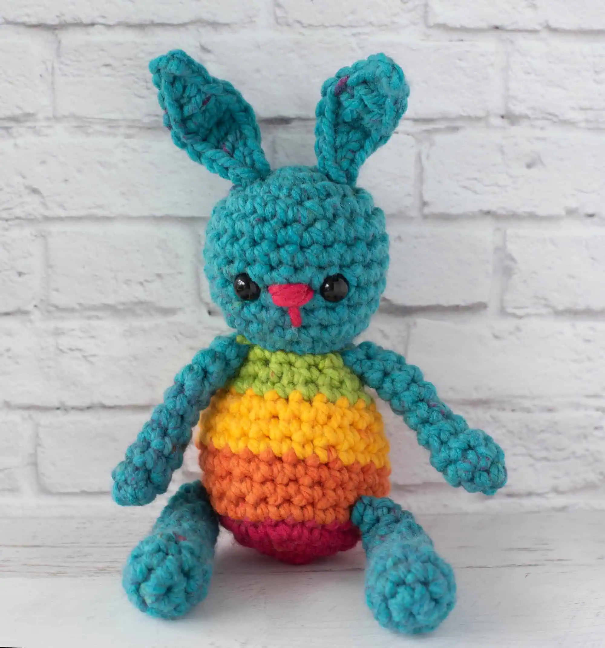 Blue crochet bunny with a rainbow striped body sitting in front of brick wall
