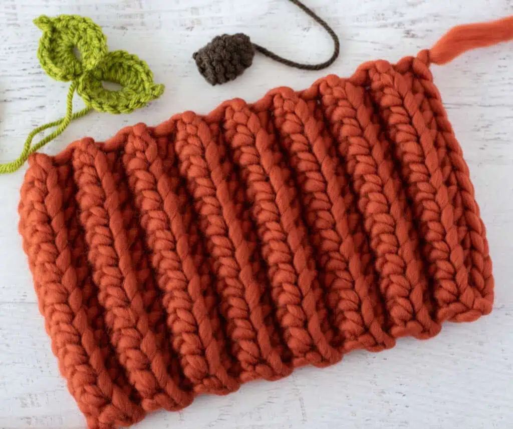 orange crochet rectangle, green crochet leaves and brown crochet stem on white wood table