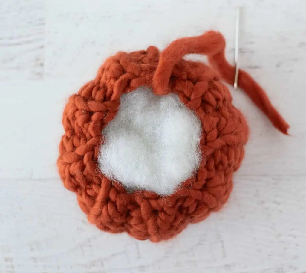 Crochet orange pumpkin in the process of stuffing, shown with white stuffing coming out of top of pumpkin and yarn threaded on a needle