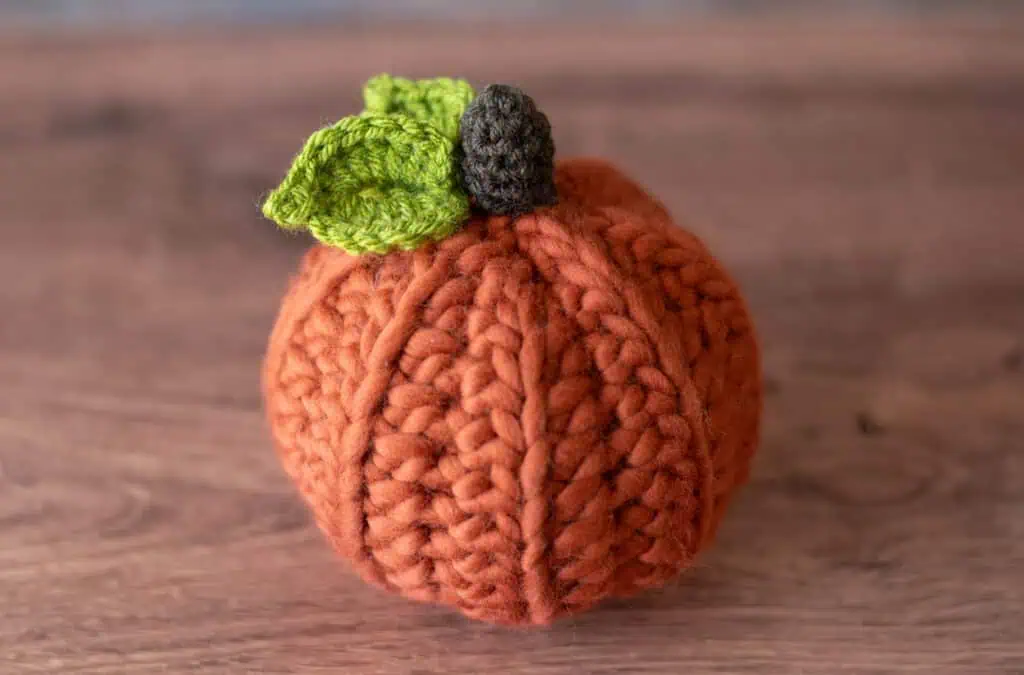 Orange crochet pumpkin with green leaves and dark brown stem on wood table